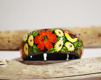 May include: A black wooden bangle with a floral design painted on it. The flowers are red, yellow, and white, and the leaves are green. The bangle is on a wooden surface.