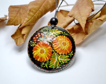 Puede incluir: Un collar con un colgante de madera negro pintado a mano con un diseño floral. El colgante presenta flores naranjas, amarillas y verdes con bayas rojas sobre un fondo negro.