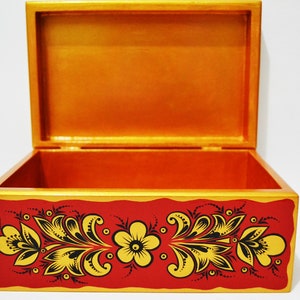 May include: Open wooden box with a red exterior and gold trim. The box is decorated with a floral design in yellow and black. The interior of the box is orange.