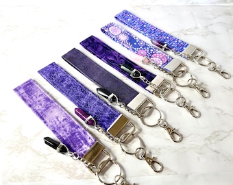 May include: Six wrist keychains in shades of purple, each with a different fabric pattern. The keychains have silver-colored hardware, including a split ring and a lobster clasp. Each keychain features a crystal pendant.