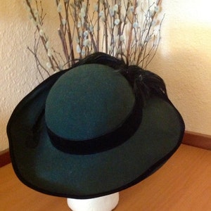 May include: A dark green felt hat with a wide brim and a black velvet ribbon. The hat has a small feather accent on the side.