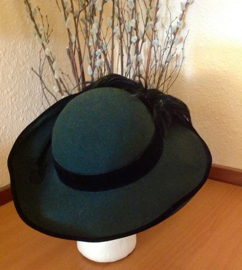 May include: A dark green felt hat with a wide brim and a black velvet ribbon. The hat has a small feather accent on the side.
