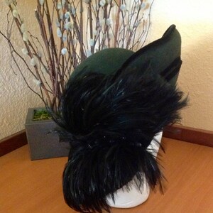 May include: A dark green felt hat with a large black feather plume. The hat is angled to the side and the feather plume is flowing down the side of the hat.