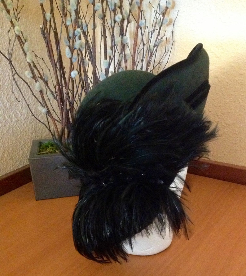 May include: A dark green felt hat with a large black feather plume. The hat is angled to the side and the feather plume is flowing down the side of the hat.