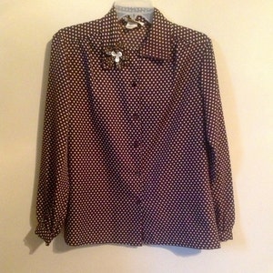 May include: A brown and white polka dot button-up shirt with a long sleeve. The shirt has a white collar and a small white flower brooch on the left side of the collar.