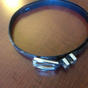 Op de afbeelding: Een zwarte leren riem met een slangenhuidpatroon en een zilveren gesp. De gesp heeft een decoratief ontwerp en de riem heeft het woord "CHINA" erop gedrukt.