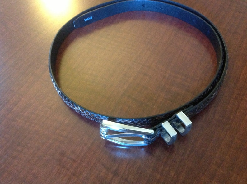 Op de afbeelding: Een zwarte leren riem met een slangenhuidpatroon en een zilveren gesp. De gesp heeft een decoratief ontwerp en de riem heeft het woord "CHINA" erop gedrukt.