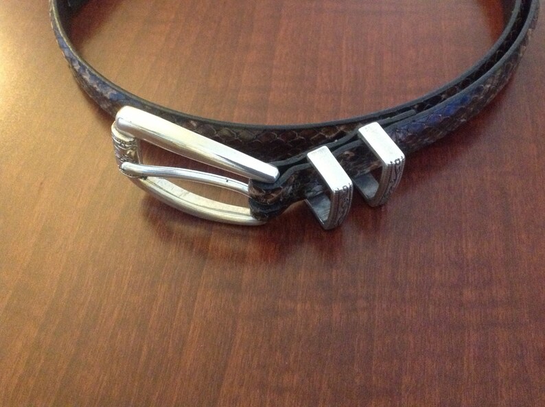 Op de afbeelding: Een bruine leren riem met een zilveren gesp en twee zilveren lussen. De riem heeft een slangenhuidpatroon.