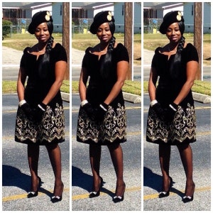 May include: A woman is wearing a black velvet dress with gold sequins. She is also wearing a black hat with a gold embellishment. She is standing in front of a green bush.