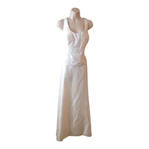 Puede incluir: Un vestido blanco largo sin mangas con escote redondo y un corpiño ajustado con intrincados bordados florales blancos. La cintura está definida por un borde festoneado que da paso a una falda fluida, lisa y sin adornos.