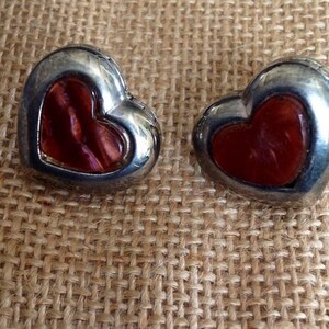 Puede incluir: Un par de pendientes de corazón plateados con piedras rojas iridiscentes, como de vidrio, en el centro.