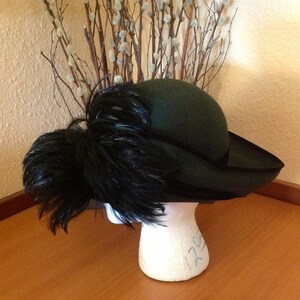 May include: A green felt hat with a wide brim and a large black feather plume. The hat is sitting on a white mannequin head.