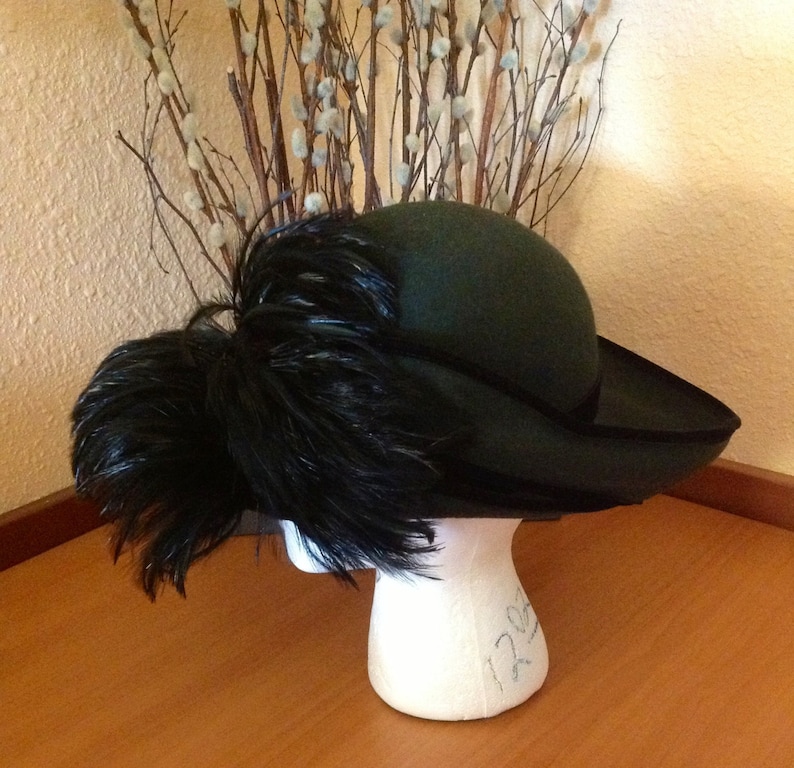 May include: A green felt hat with a wide brim and a large black feather plume. The hat is sitting on a white mannequin head.