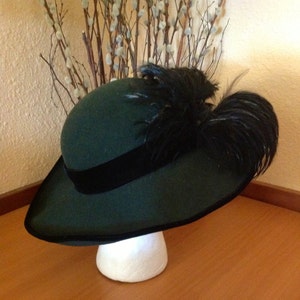 May include: A dark green felt hat with a wide brim and a black velvet band. The hat is adorned with a large black feather plume.