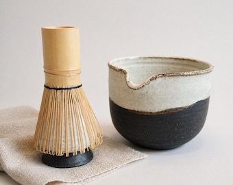 May include: A bamboo matcha whisk with a black base and a two-toned ceramic bowl featuring a white upper section and a dark charcoal lower section, both resting on a light beige textured fabric cloth.