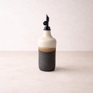 May include: A ceramic oil dispenser featuring a two-tone design with a matte black base and a cream-colored top section. The bottle is topped with a black plastic pour spout with a hinged lid, creating a functional kitchen vessel.