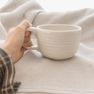 May include: A cream-colored ceramic mug with a rounded body and a handle. The mug has horizontal ridges around the exterior. The mug is resting on a beige linen cloth.