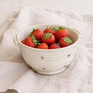 May include: A ceramic bowl filled with fresh, ripe strawberries. The bowl is off-white with small drainage holes and handles. The strawberries are bright red with green leaves, resting on a beige linen cloth.