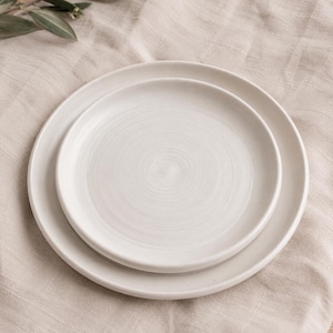 May include: Two white ceramic plates, one nested inside the other, on a beige linen surface. The plates have a simple, minimalist design with a slightly raised rim. A sprig of green leaves is visible in the upper left corner, adding a touch of natural detail.