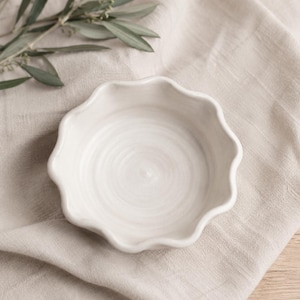 May include: A small, off-white ceramic bowl with a scalloped edge. The bowl has a circular interior with a spiral design. The bowl is on a beige linen cloth, with a sprig of green leaves in the background. The bowl is likely used for serving food.