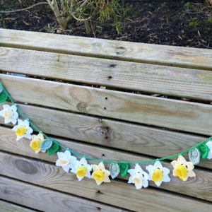 Puede incluir: Una guirnalda de crochet con narcisos blancos y amarillos y hojas verdes. La guirnalda está colgada en una valla de madera.