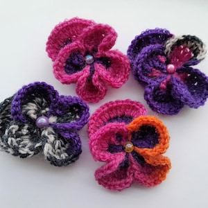 May include: Four crocheted flower embellishments in various colors, including pink, purple, black, and orange. Each flower has a small, iridescent bead in the center.