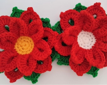 May include: Two red crocheted poinsettia flowers with green leaves. The center of one flower is yellow and the center of the other flower is white.