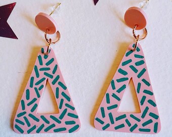 Puede incluir: Un par de pendientes triangulares de color rosa con un patrón geométrico verde. Los pendientes tienen un pequeño círculo rosa en la parte superior y un gancho dorado.