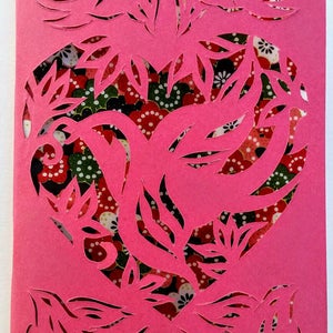 May include: A pink paper card with a heart-shaped cutout design. The cutout features intricate floral patterns and two birds. The background is a floral patterned fabric.