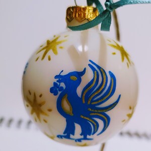 Könnte beinhalten: Weißer Weihnachtsbaumschmuck mit einem blauen und goldenen Phönix-Design. Der Phönix hält einen Zweig mit Beeren im Schnabel. Der Schmuck ist mit goldenen Sternen verziert.