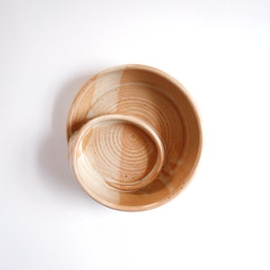 May include: A ceramic serving dish with two sections. The larger section is a round bowl with a white glaze and brown speckles. The smaller section is a round bowl with a brown glaze and white speckles. The bowls are connected by a curved handle.