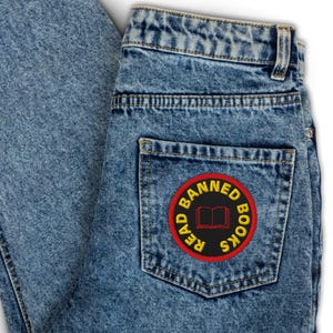 May include: A pair of blue denim jeans with a red and yellow embroidered patch on the back pocket. The patch says "Read Banned Books" with an open book icon in the center.