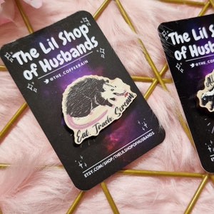May include: Wooden pin featuring a cartoon opossum with the text "Eat Trash, Scream." The pin is on a black card with the shop name "The Lil Shop of Husbands" and the Etsy shop URL.