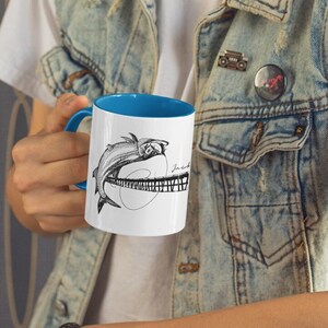 May include: White ceramic mug with a blue interior, featuring a black and white illustration of a fish leaping over a bridge. The mug has a curved handle and is held by a person. The background includes a decorative wall and a ram head.
