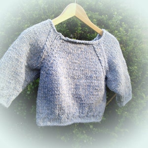 May include: A light blue knitted sweater with long sleeves hangs on a wooden hanger. The sweater has a round neckline and a textured knit pattern. The garment is displayed against a backdrop of green foliage.