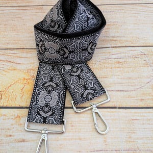 May include: Black and white patterned bag strap with silver-colored metal clasps. The strap features a repeating paisley-like design in white against a black background. The strap is folded over itself, showing the pattern on both sides.