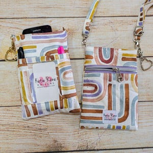 May include: Two small crossbody bags with a white background and a colorful abstract pattern of arches and lines in shades of brown, purple, and blue. The bags have a zipper closure and a small business card holder with the text "Katie Rene Designs" and a website address.