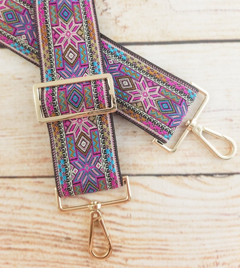 May include: A pair of colorful patterned fabric straps with gold metal clasps and swivel hooks. The straps have a geometric design with pink, blue, and gold accents.