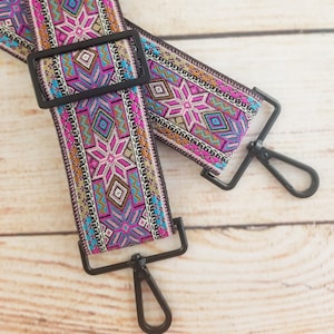 May include: A pink, blue, and gold patterned woven guitar strap with black metal clasps.