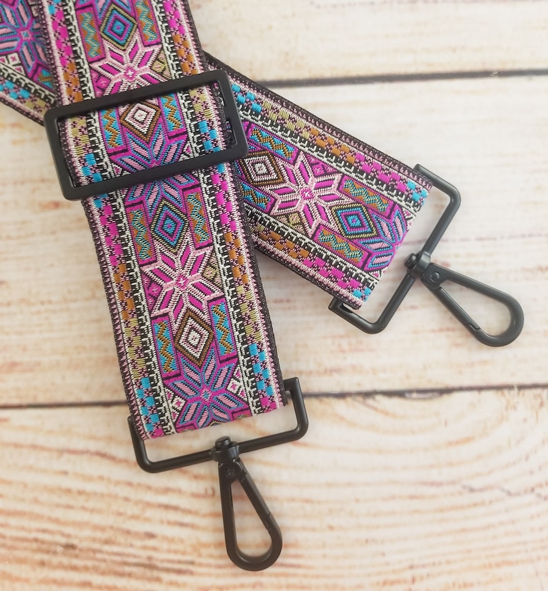May include: A pink, blue, and gold patterned woven guitar strap with black metal clasps.