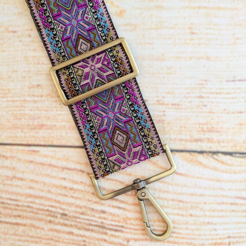 May include: A close-up of a colorful woven strap with a gold metal buckle and a swivel clasp. The strap has a geometric pattern in shades of pink, blue, and brown.