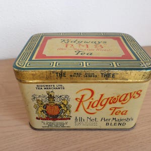 Può includere: Scatola di tè Ridgways vintage, di forma rettangolare con coperchio decorativo. La scatola presenta testo e grafica in rosso, oro e verde, tra cui le parole "Ridgways Tea" e "Her Majesty's Blend". La scatola è color crema.