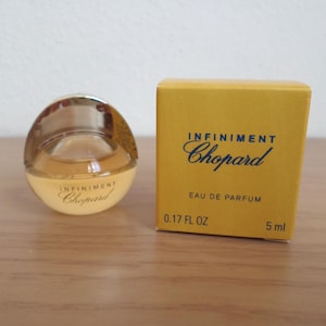 Könnte beinhalten: Ein kleiner, runder Parfümflakon und eine gelbe Schachtel. Die Flasche ist klar mit einem goldfarbenen Verschluss und enthält eine hellbernsteinfarbene Flüssigkeit. Auf der Schachtel stehen die Worte "INFINIMENT Chopard" und "EAU DE PARFUM". Die Flasche ist 5 ml.
