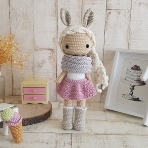 Puede incluir: Una muñeca de crochet con orejas de conejo, una bufanda gris, una falda rosa y botas grises. La muñeca tiene el pelo largo y rubio y está de pie sobre una superficie de madera.
