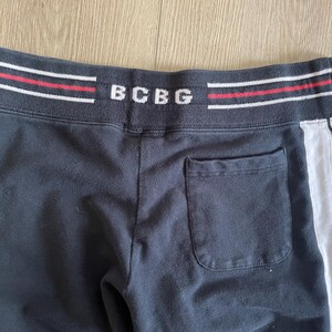 May include: Black sweatpants featuring a wide elastic waistband with the text BCBG in white, accented by horizontal red and white stripes. The pants include a single back pocket and a vertical white side panel with thin black pinstripes.