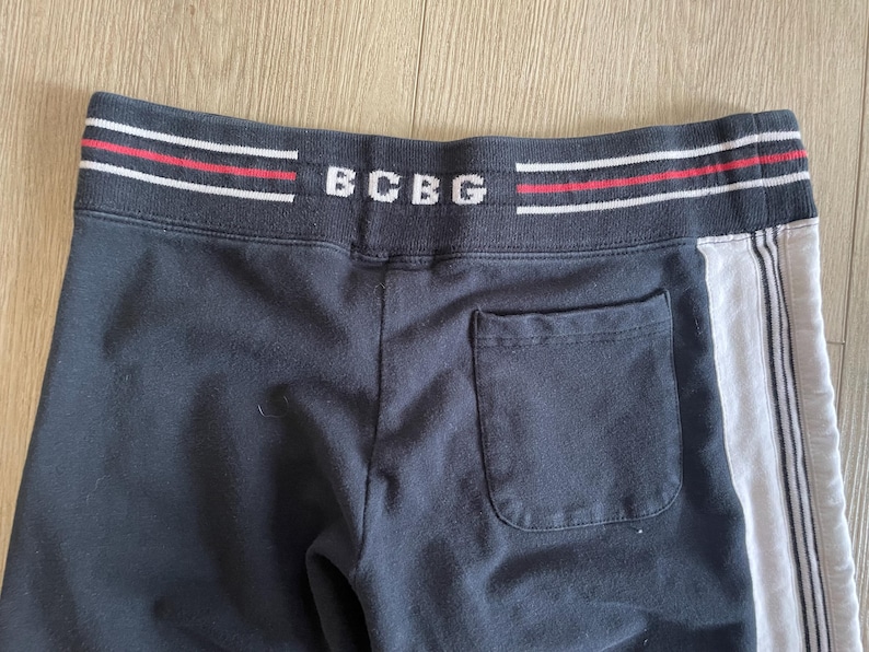 May include: Black sweatpants featuring a wide elastic waistband with the text BCBG in white, accented by horizontal red and white stripes. The pants include a single back pocket and a vertical white side panel with thin black pinstripes.