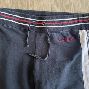 May include: Black sweatpants featuring a ribbed waistband with red and white stripes and a drawstring. The text BCBG is embroidered in red on the front. A white vertical side panel with thin black stripes runs down the side of the garment.