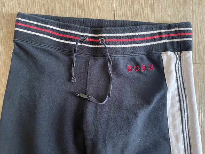 May include: Black sweatpants featuring a ribbed waistband with red and white stripes and a drawstring. The text BCBG is embroidered in red on the front. A white vertical side panel with thin black stripes runs down the side of the garment.