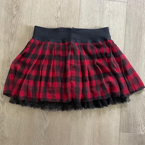 May include: A red and black plaid pleated skirt with a black waistband and a black lace trim at the hem. The skirt is made of a lightweight fabric and is designed to be worn at the waist.