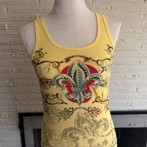May include: A yellow ribbed tank top featuring a central fleur-de-lis graphic embellished with small silver studs. The design includes a red circular background behind the fleur-de-lis, surrounded by black and grey scrollwork patterns.